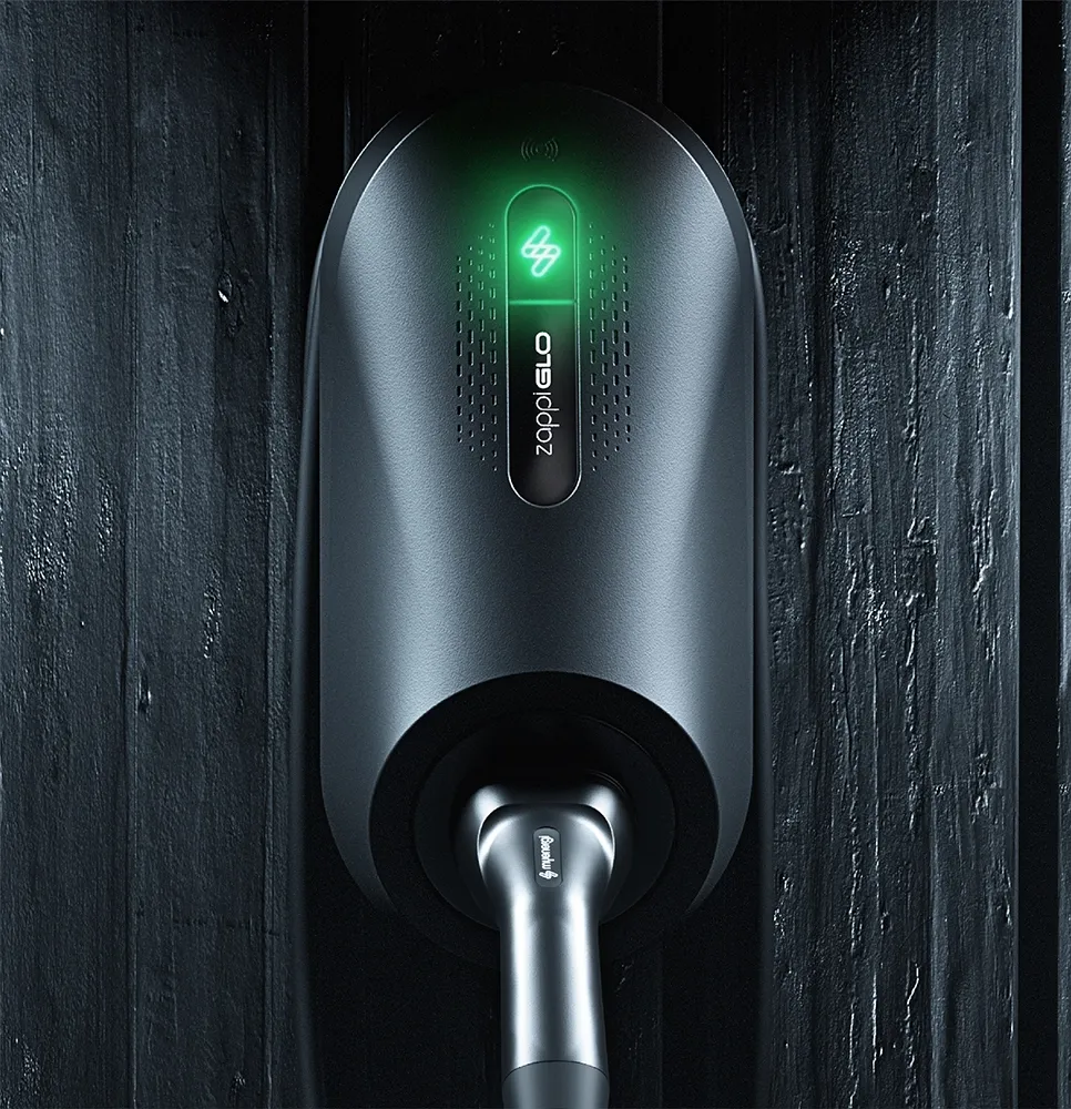 Front-facing view of a black myenergi Zappi GLO EV charger mounted on a dark timber-clad wall. The charger is illuminated with a soft green LED light at the top, displaying the myenergi logo, and has a tethered charging cable plugged into the socket below.